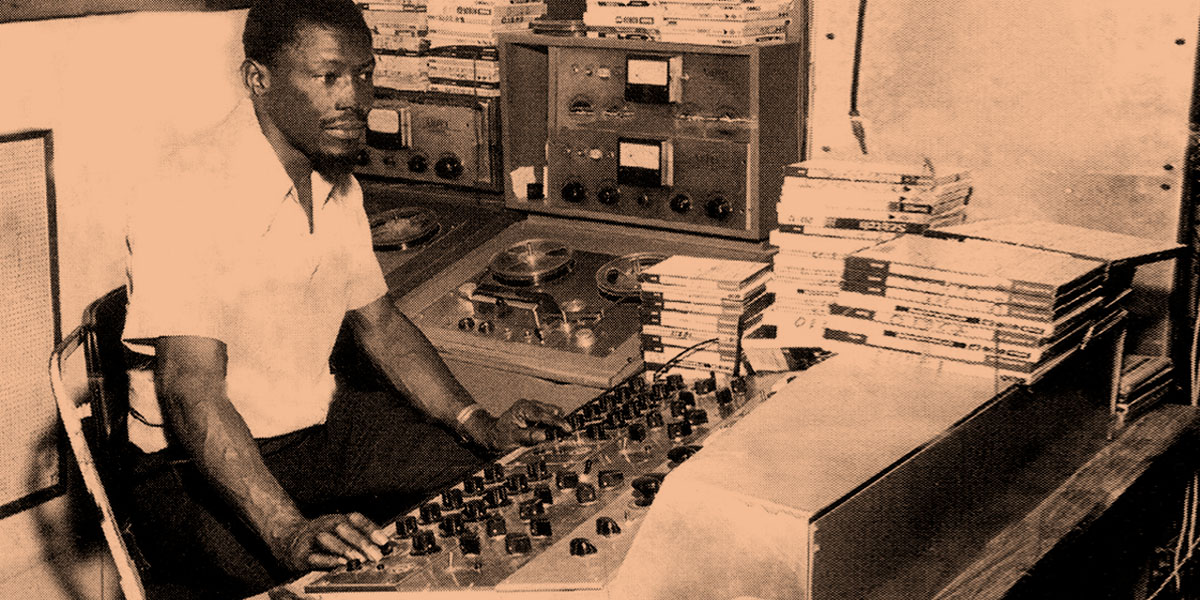 Coxsone Dodd at the control panel in Studio One, surrounded by recording equipment and stacks of records, showcasing his role as a legendary producer and artist scout in Jamaican music history.