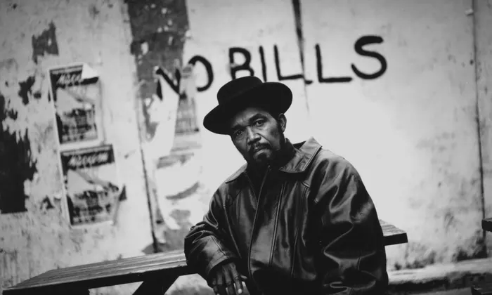 Prince Buster, the legendary Jamaican musician and producer, posing in front of a 'No Bills' graffiti wall, exuding a stern and contemplative demeanor. Photo captures his influential presence in the reggae and ska music scenes.